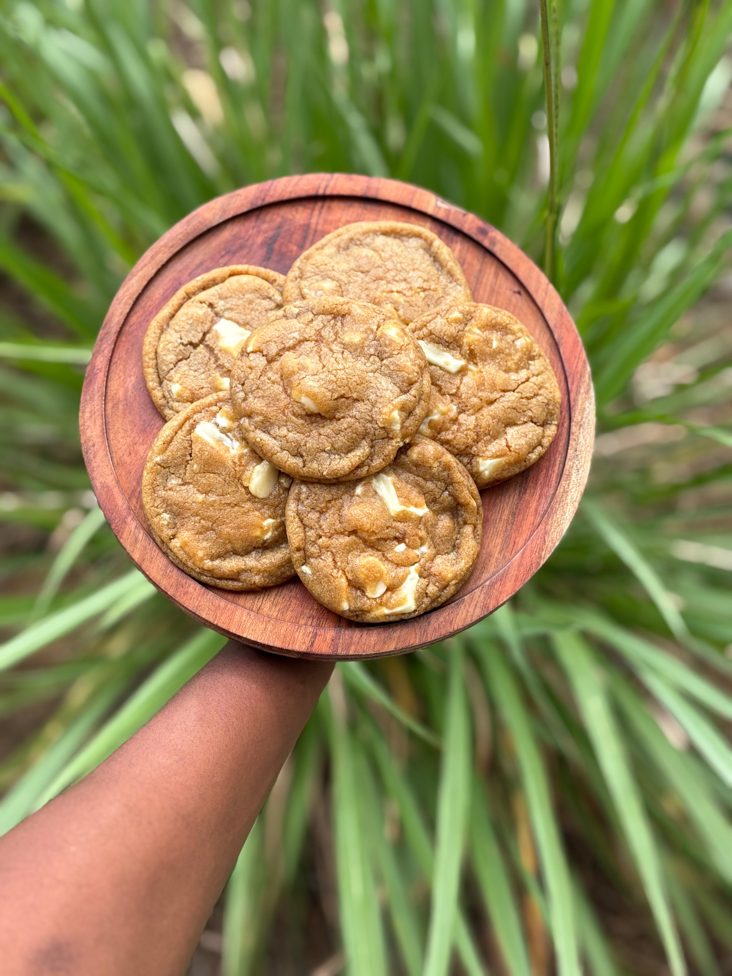 Six white chocolate chunk cookies arranged on a round wooden board, held up outdoors against tall green grasses.