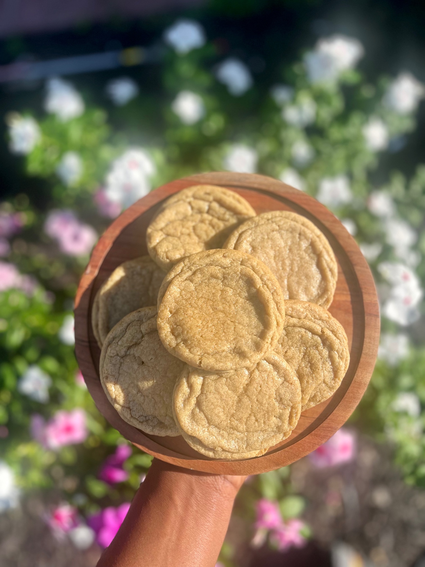 A wooden board stacked with soft golden sugar cookies, held up in dappled sunlight in front of a flowering garden.